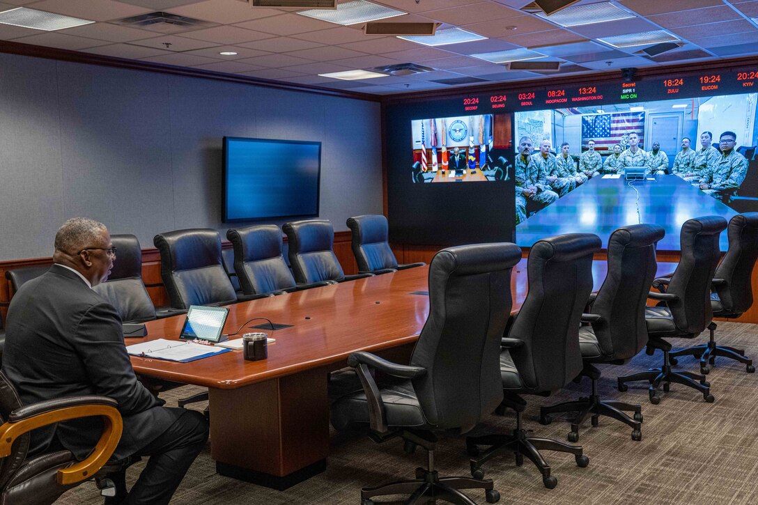Secretary of Defense Lloyd J. Austin III sits at a large conference room table while talking to troops on large monitors at the other end.