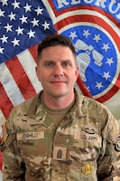 U.S. Army Soldier standing in front of the American Flag and a Recruiting Bell Flag.