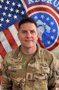 U.S. Army Soldier standing in front of the American Flag and a Recruiting Bell Flag.