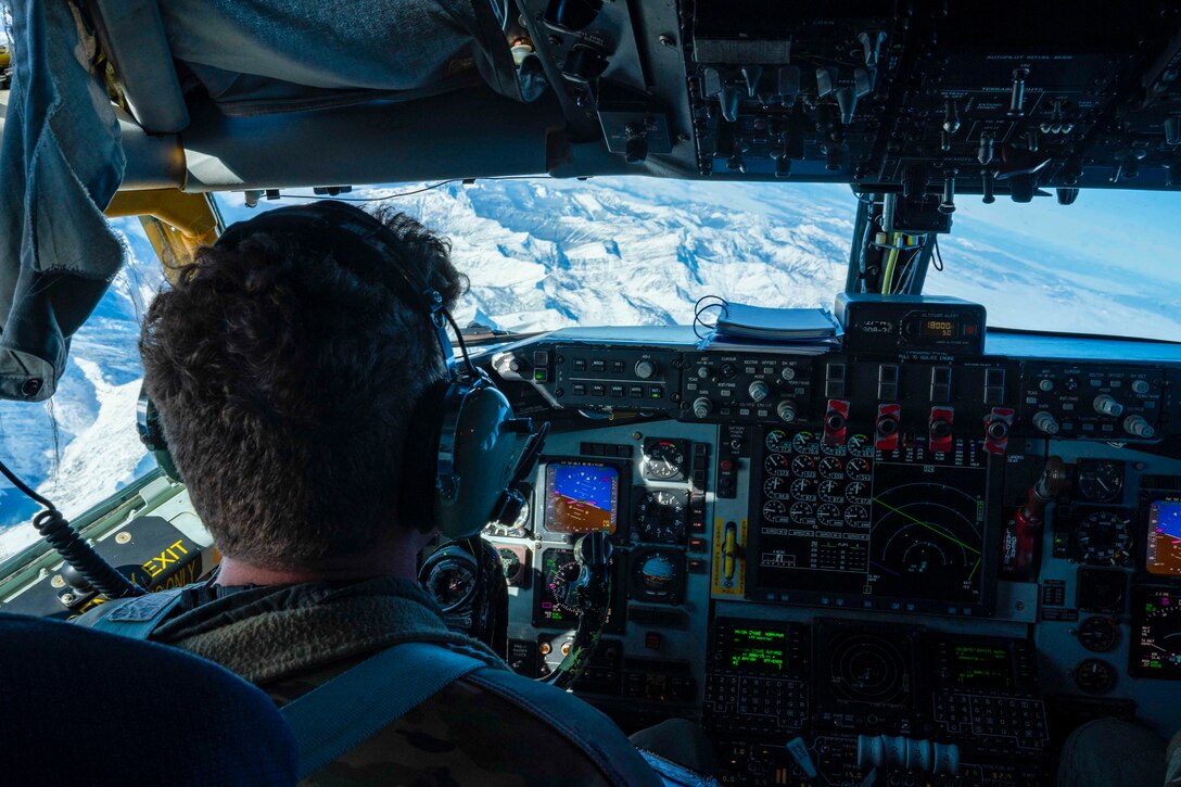 An airman sits in the cockpit of an airborne aircraft looking out over a mountain.