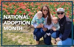A family of four is posed in front of a field of pink flowers. The words National Adoption Month appear on the left side of the photo. On the right side is Health Services Technician 1st Class Petty Officer Jessica Hulon, and her spouse, Amber, is posed beside her. Both Amber, and Jessica are holding one of their respective children on their knee. The kids names are Leo and Penelope.
