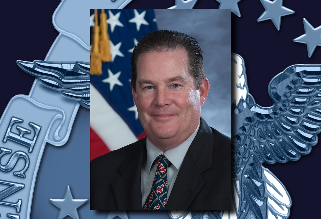 A headshot of a fair skinned man with a dark suit on and a US Flag behind him.