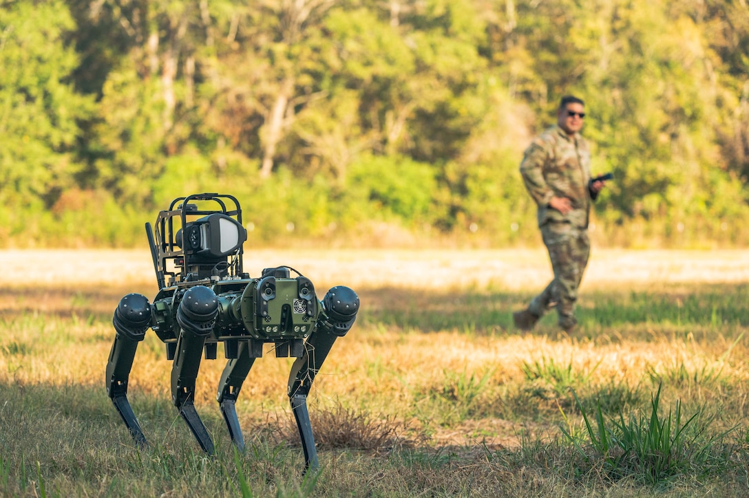 A robot stands on the grass as an airman observes in the background.