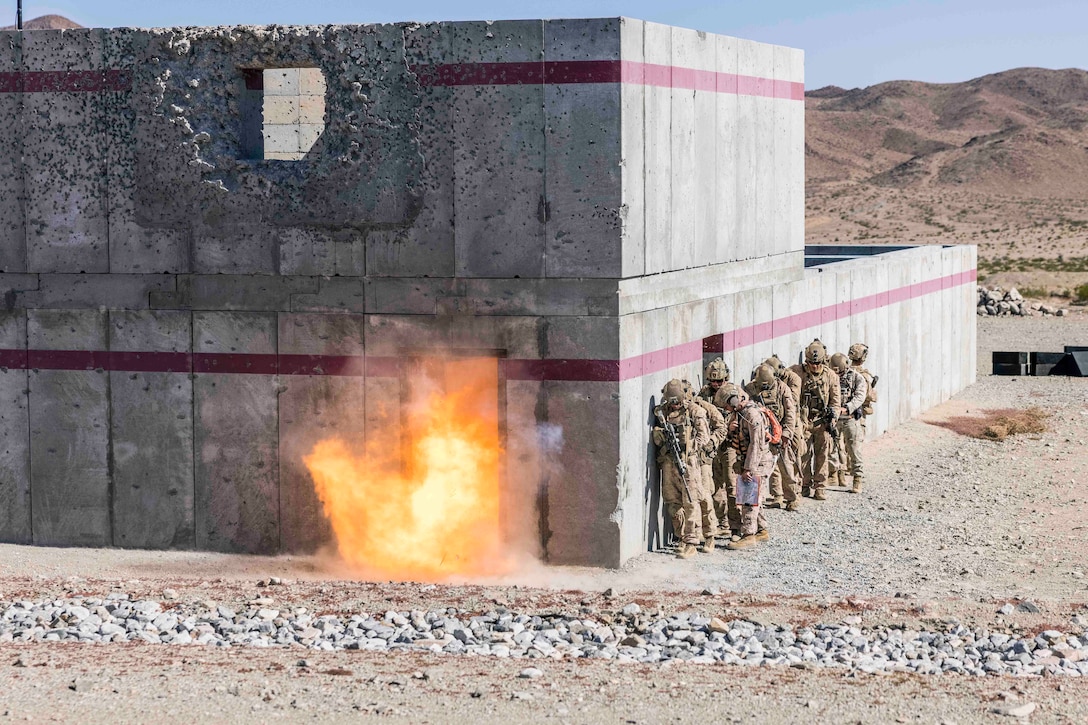 Marines stand in a line on the side of a building as an explosive detonates in a door frame.
