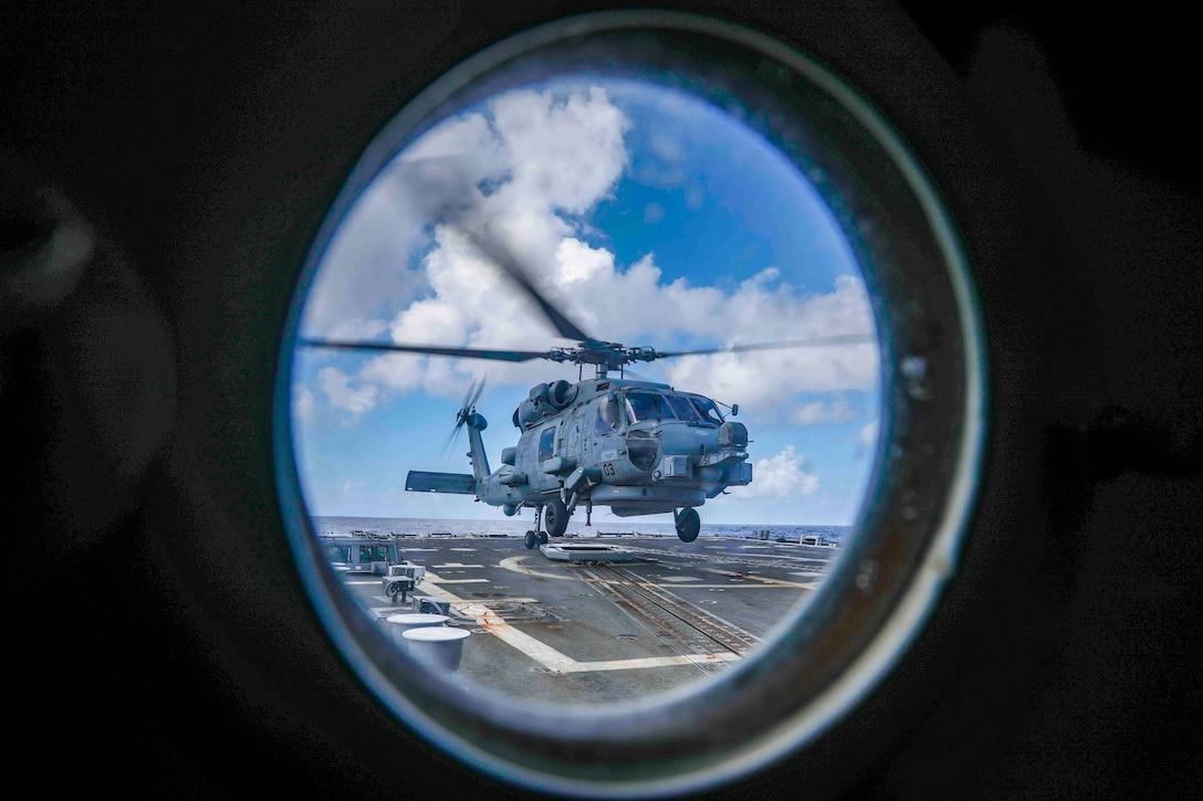 An aircraft seen through a porthole hovers over the deck of a ship.