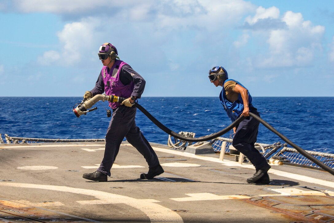 Two sailors carry a fuel hose aboard a ship with a body of water in the background.