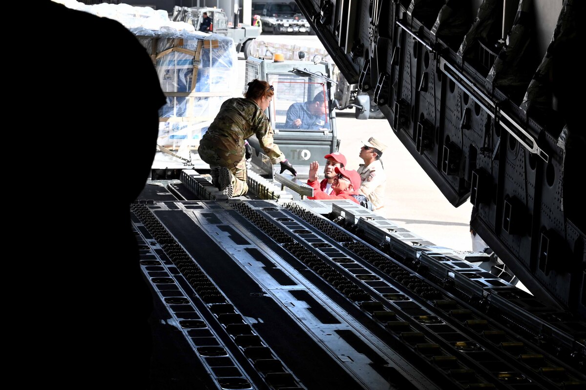 A female airman leans forward to talk to men loading cargo into an aircraft.