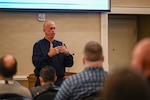 A man in a blue military jacket addresses a crowd in front of him.