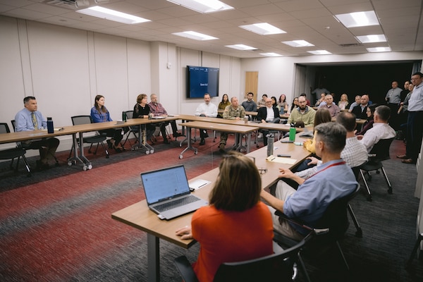 People sit at tables set up in U-shape around a conference room.