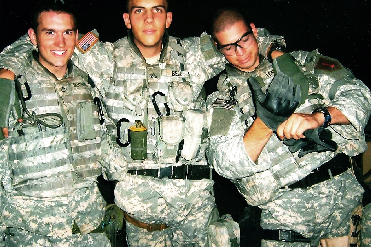 Three men in combat gear pose for a photo with the man in the middle wrapping his arms around the other two.