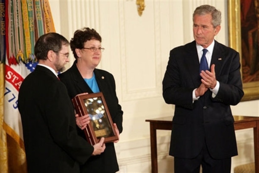 A man claps as two others hold up a box that contains a medal.