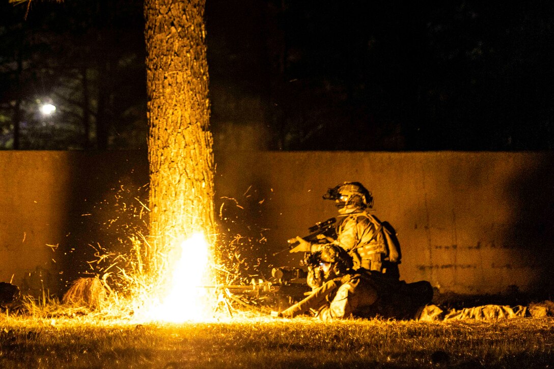 A Marine lies on the ground firing a weapon while another Marine kneels beside at night.