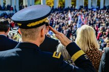An Army general has his back to the camera and is saluting. There are hundreds of people in the distance who are participating in the ceremony.