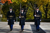 Three members of the Army's Old Guard unit are marching toward the photographer. They are wearing dark overcoats, hats and sunglasses, and the two on the right are carrying rifles on their right shoulders.