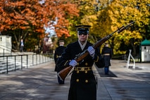 A female member of Army's Old Guard unit is marching in front of the Tomb of the Unknown Soldier while holding a rifle in front of her at a 45 degree angle. Behind her are trees with orange and yellow leaves.