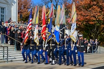 Military members are carrying the flags for each branch of the military and the U.S. flag while marching to their positions near the Tomb of the Unknown Soldier. Behind them is a large crowd of people watching the ceremony.