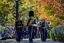 Members of The U.S. Army Band are marching toward the photographer between a row of wreaths and a row of bushes. Some are carrying various instruments, and the band member in the middle of the picture is wearing a tall, fuzzy hat and has a red sash across his chest and he is carrying a large silver mace in one hand.