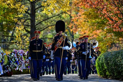 Members of The U.S. Army Band are marching toward the photographer between a row of wreaths and a row of bushes. Some are carrying various instruments, and the band member in the middle of the picture is wearing a tall, fuzzy hat and has a red sash across his chest and he is carrying a large silver mace in one hand.