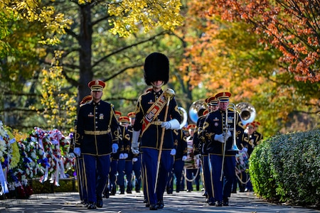 Members of The U.S. Army Band are marching toward the photographer between a row of wreaths and a row of bushes. Some are carrying various instruments, and the band member in the middle of the picture is wearing a tall, fuzzy hat and has a red sash across his chest and he is carrying a large silver mace in one hand.