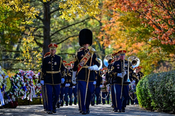 Members of The U.S. Army Band are marching between a row of wreaths and a row of bushes. Some are carrying instruments, and the band member in the middle is wearing a hat, a red sash across his chest, and he is carrying a large mace in one hand.