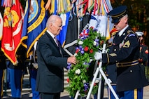 President Biden is placing ceremonial wreath onto a white metal stand with the assistance of an Army soldier who is dressed in a dark blue uniform with hat.