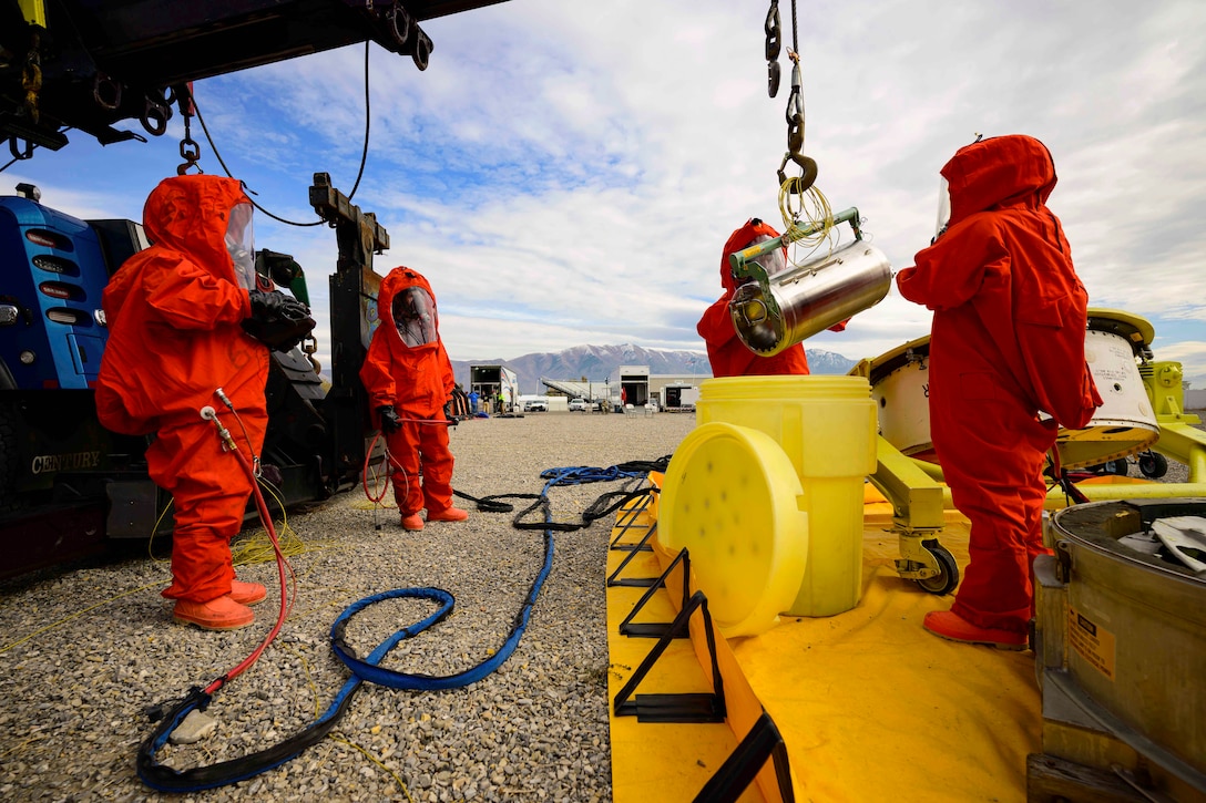 Four people wearing hazmat suits handle a canister at a work site with mountains in the background.