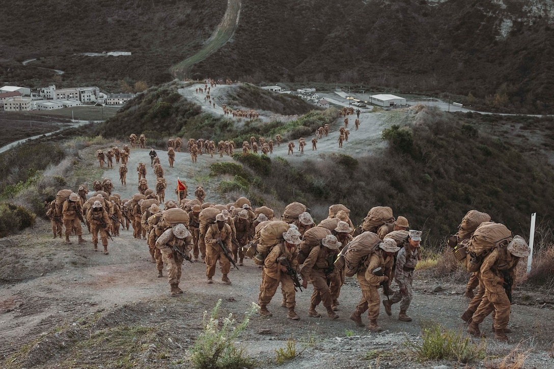 Dozens of Marines carrying sacks on their backs and weapons hike a dirt road in the mountains with buildings in the distance.