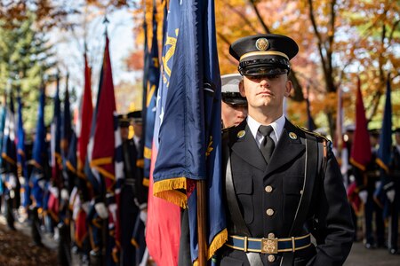 An Army Soldier stands at attention holding a flag from one of the US states. He is in front of a long row of other service members who are each holding another US state flag.