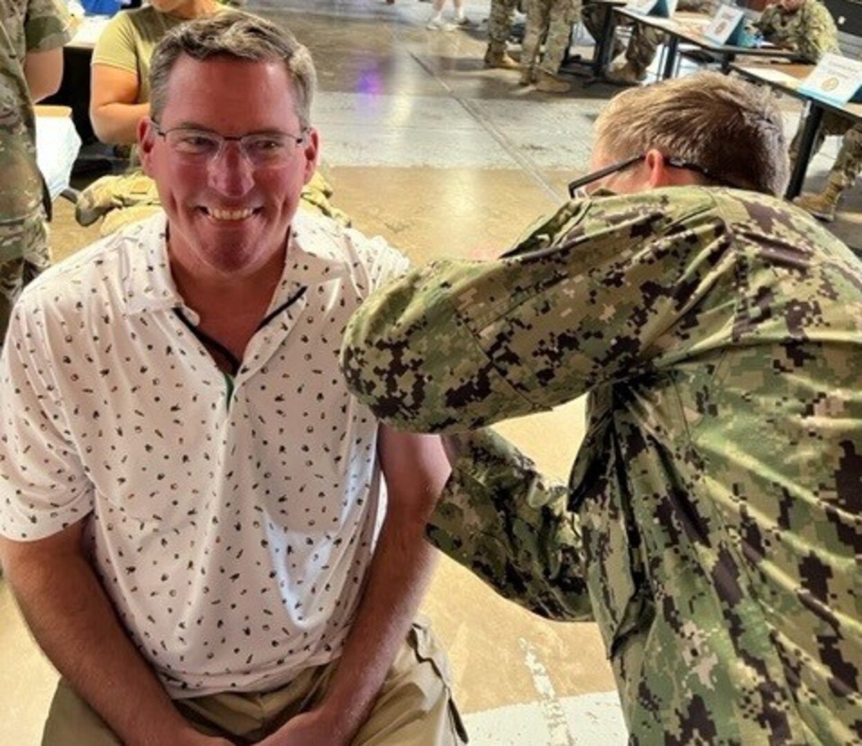 Man in white shirt sits as man on right in uniform administers flu shot.