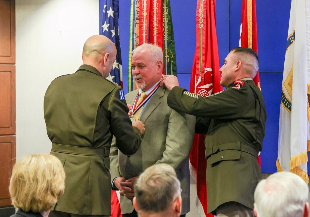 Man in suit in front of US and USACE flags receives medal around his neck from 2 uniformed Army officers.