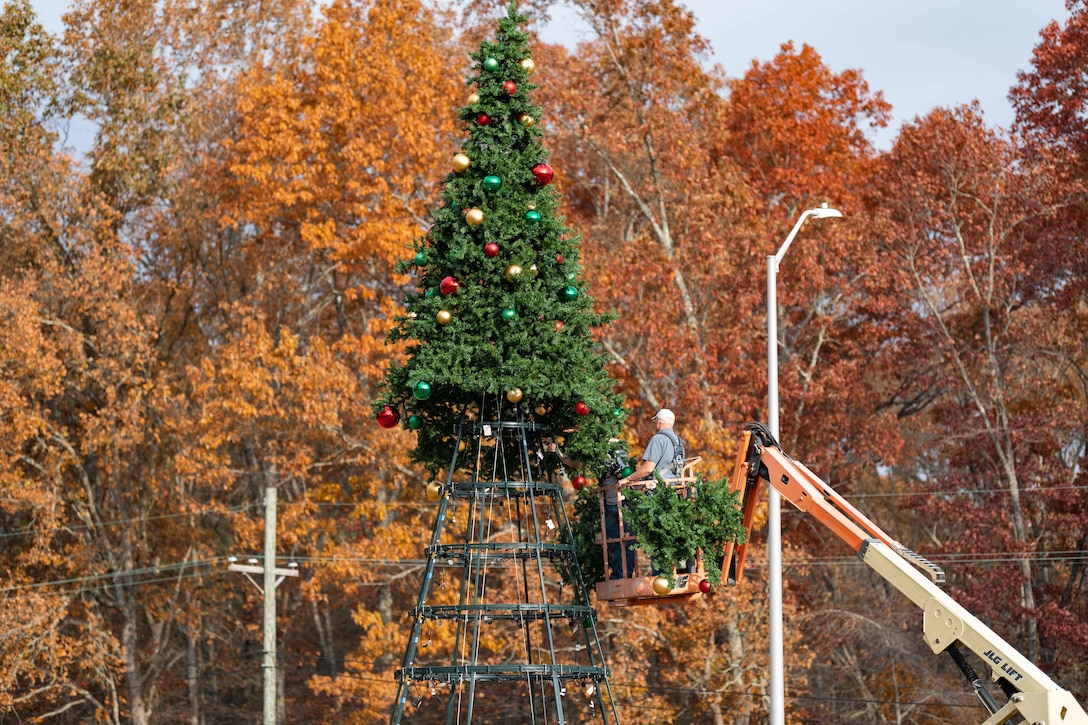 A person in a cherry picker assembles a large Christmas tree with fall foliage in the background.
