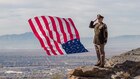 U.S. Army Soldier stands on a cliff with the American Flag blowing behind him.