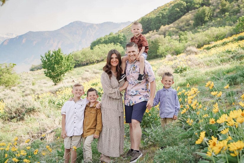 Husband and wife stand on a grassy hill with their four sons. One son sits on his father's shoulders, one son is holding his father's hand, and two stand arm in arm beside their mother.