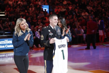 U.S. Army Soldier stands on an NBA court holding a personalized jersey. Two NBA cheerleaders stand on either side of him