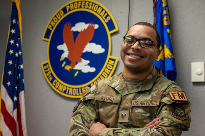 Airman stands with his arms crossed In front of the 375th Comptroller Squadron emblem.