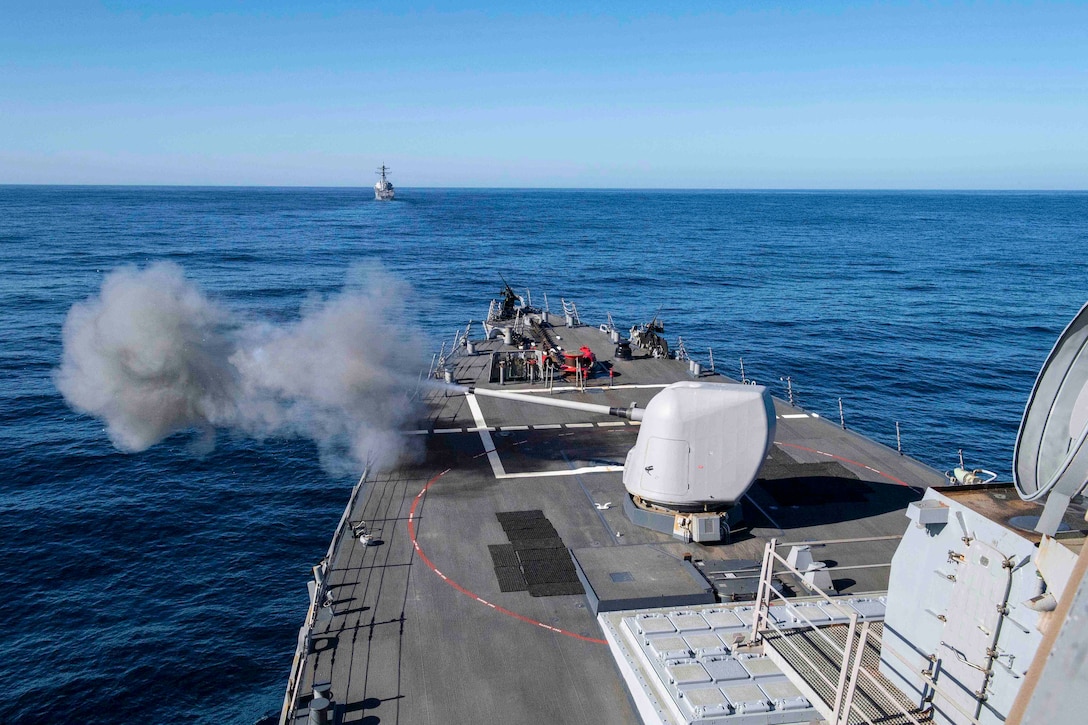 A weapon fires from the deck of a ship traveling through a body of water.