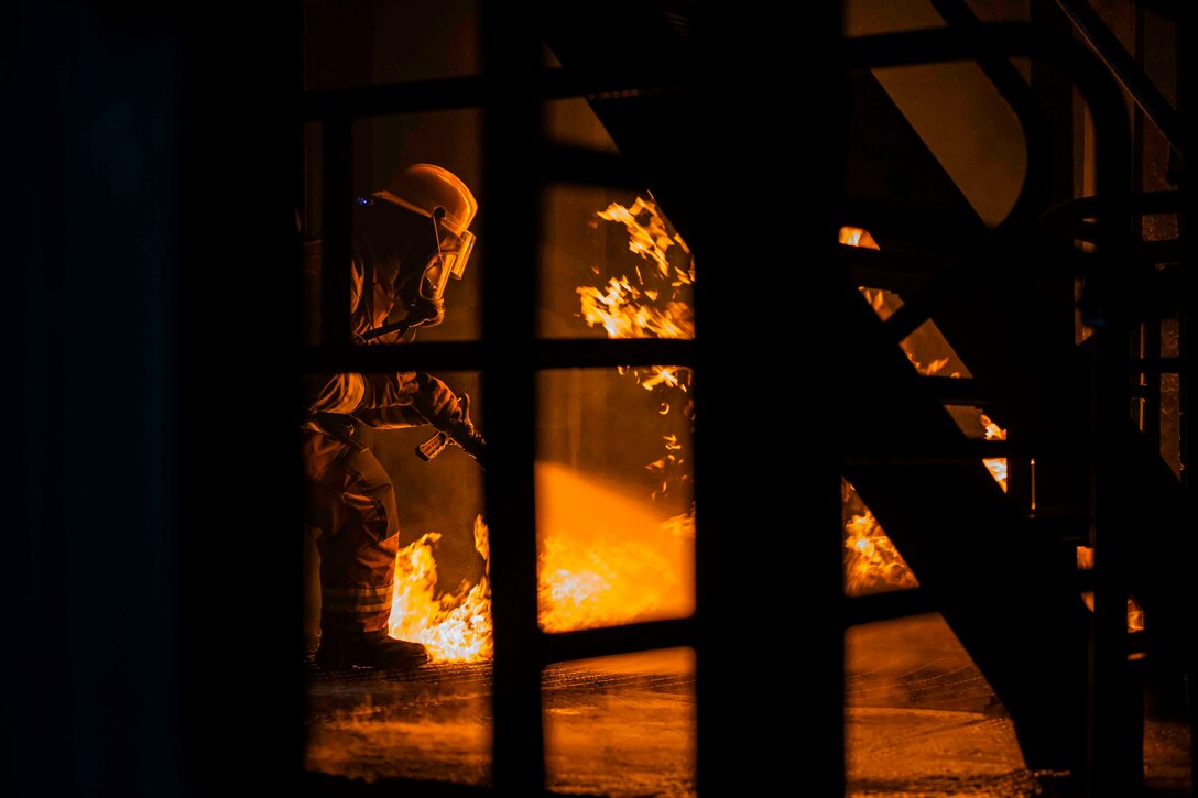 A person in fire protective gear uses a hose to spray water on a fire inside a structure.