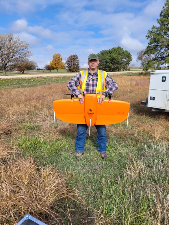 A man in a blue plaid shirt with a tan hat holds an orange airplane object while standing in grass.