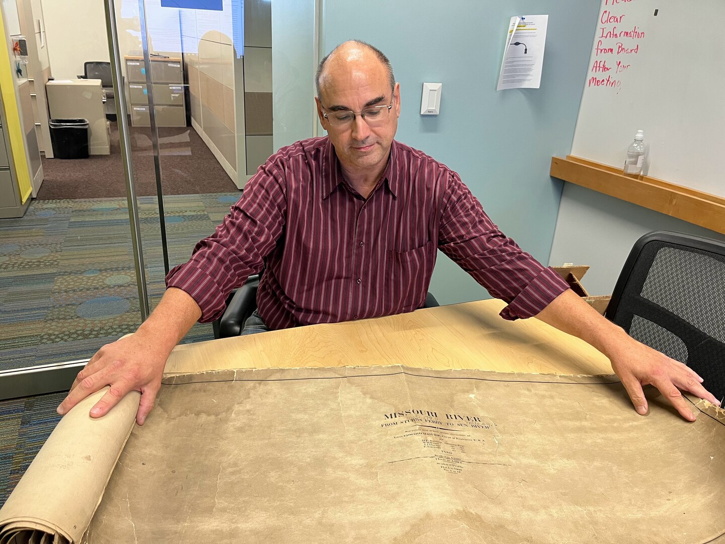 A man in a red shirt unrolls a brown sheet of paper and gazes down at it.