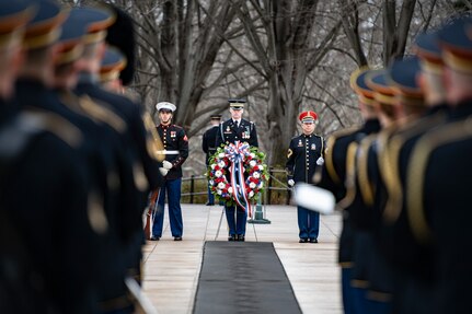 Three service members are visible in the distance: one is holding a rifle against the ground, one is behind a wreath and one is holding a bugle. In front of them is a black rubber mat that sentinels of the Tomb of the Unknown Soldier step across as they guard the tomb.