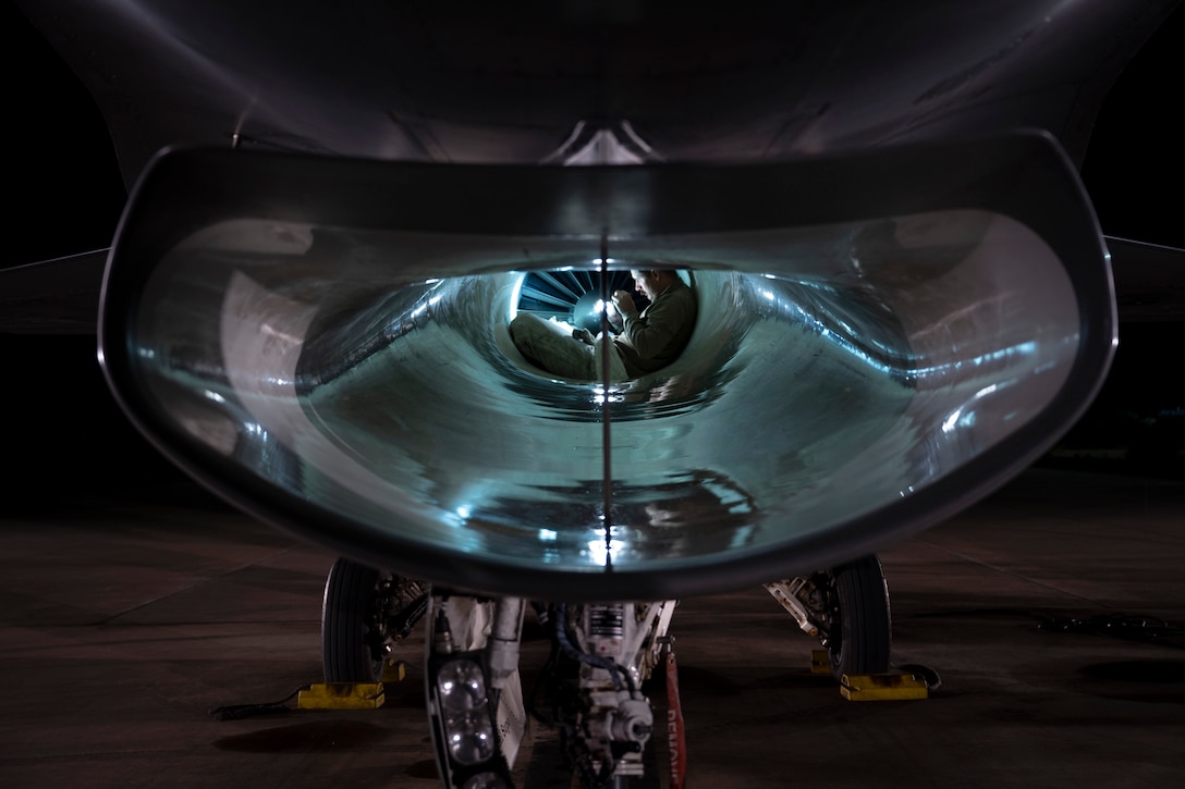 An airman shines a flashlight on an aircraft's engine as seen through an open port.