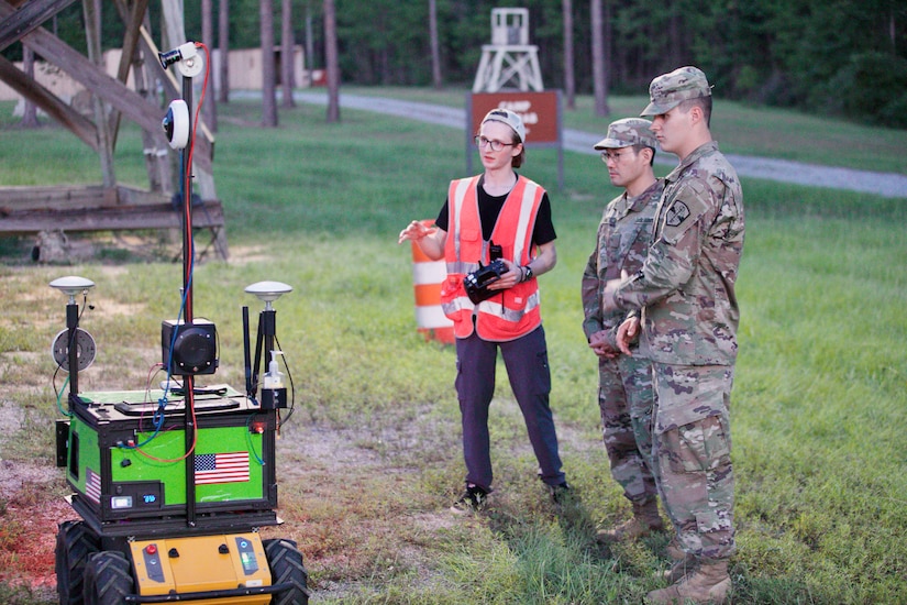 Two soldiers in combat uniform stand next to a man wearing a reflective vest. They observe a green robot that is mounted on a wheeled yellow chassis.