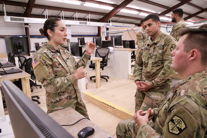 Three Soldiers stand together in a large room filled with computer screens. They are shown talking to each other.