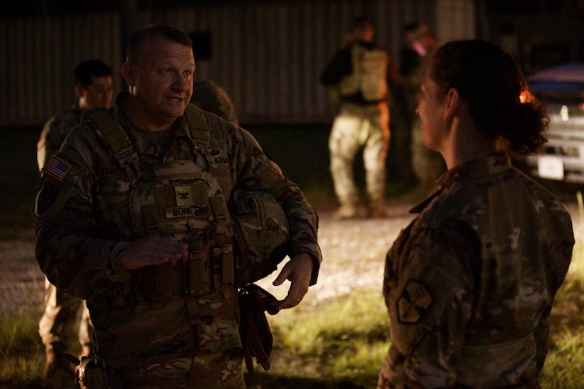 Two Soldiers in combat uniforms stand together in the dark speaking. They are illuminated by lighting coming from a vehicle in the background.