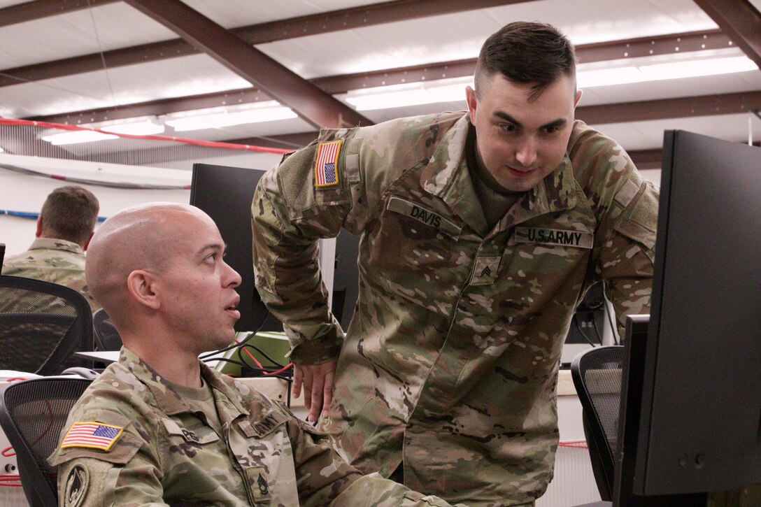 Two Soldiers work speak while looking at a computer screen in a large room.