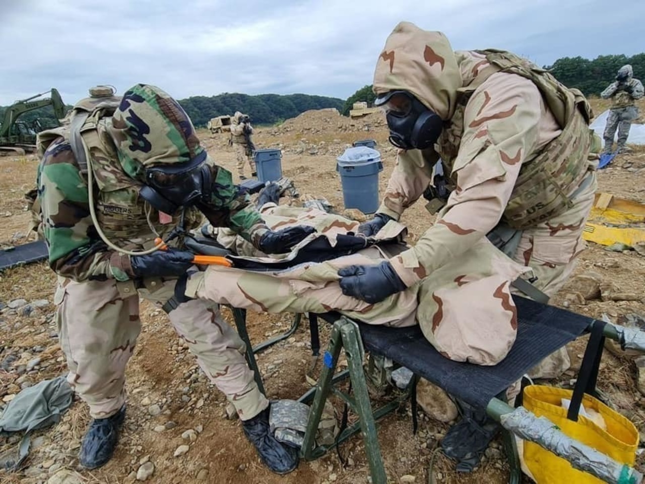 Soldiers wearing gas masks and protective suits work on a simulated casualty.