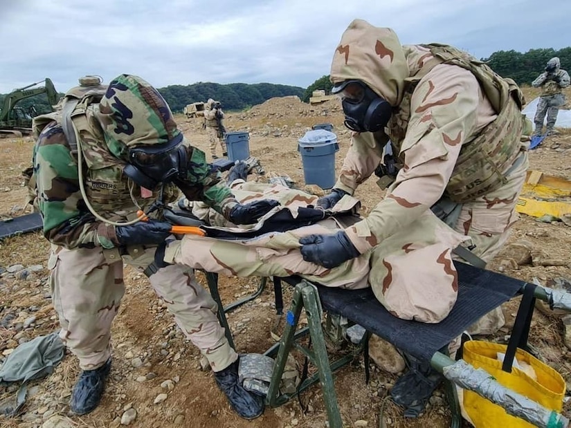 Soldiers wearing gas masks and protective suits work on a simulated casualty.