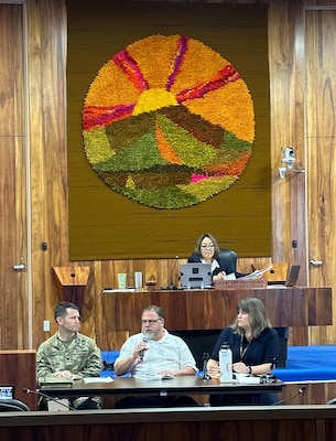 Three people sit at a table on a stage and address a council meeting. On a higher stage behind them a woman presides over the meeting.