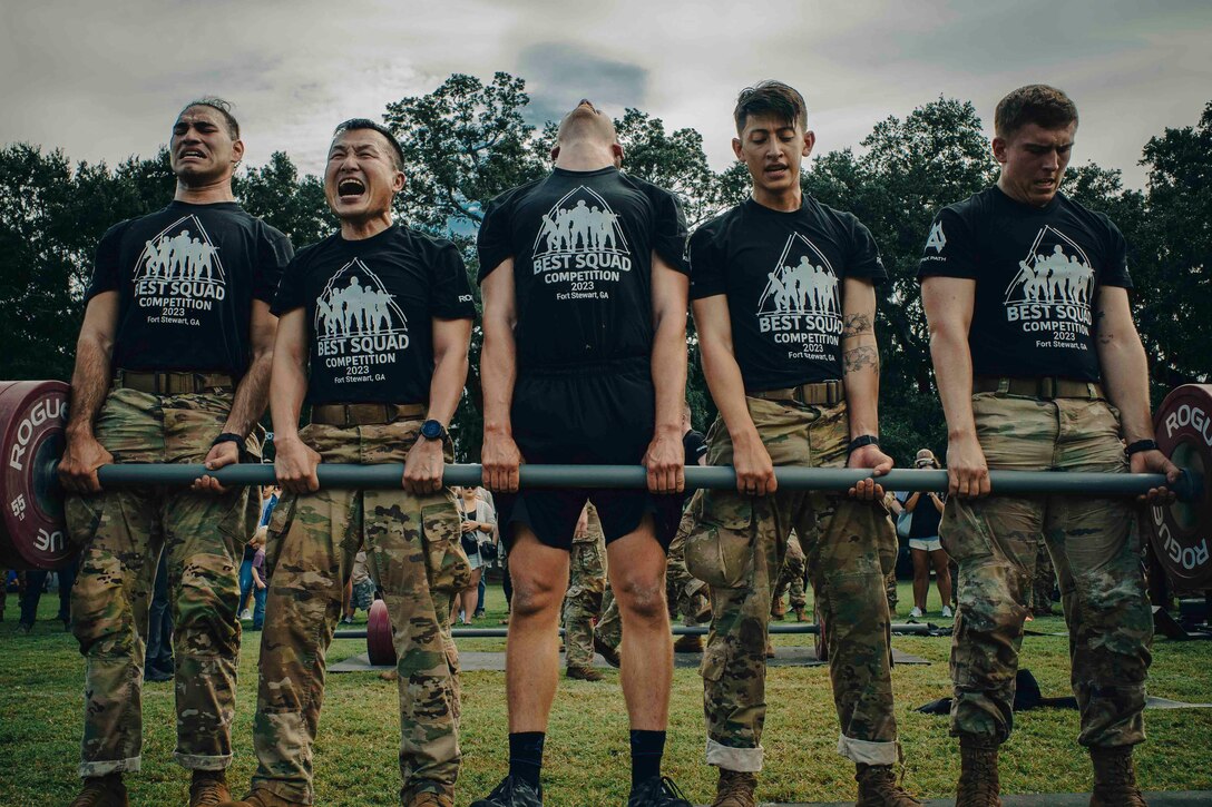 Five soldiers hold onto a barbell while some grimace.