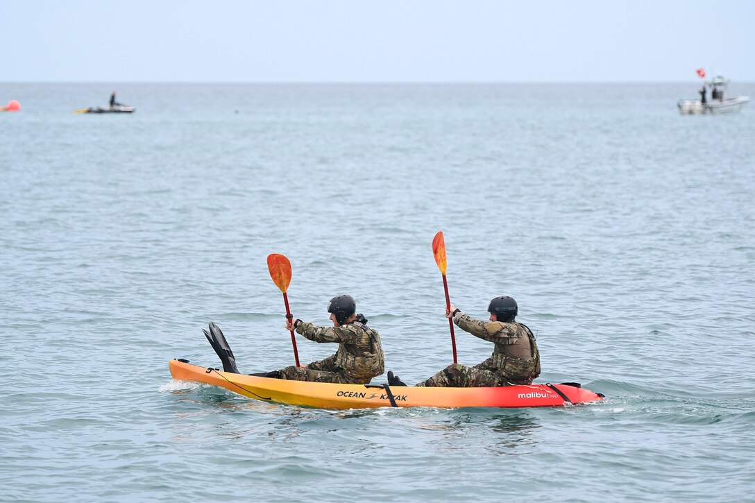 Two service members use oars to paddle a kayak while wearing flippers with a small boat in a blurred background.
